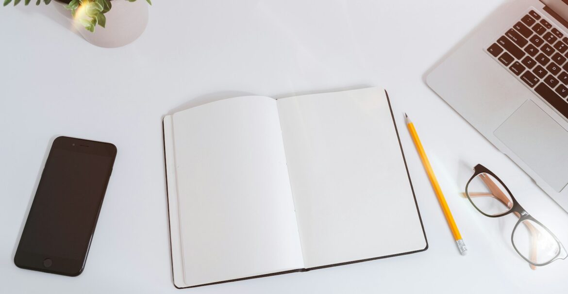 An open empty notebook on a white desk next to an iPhone and a MacBook